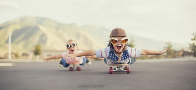 A young boy and girl are wearing flying goggles while outstretching their arms to attempt flying while on skateboards. They have large smiles and are imagining taking off into the sky.